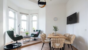 Un salon moderne avec une table à manger en bois, des chaises vertes et beiges, un téléviseur fixé au mur et de grandes fenêtres.