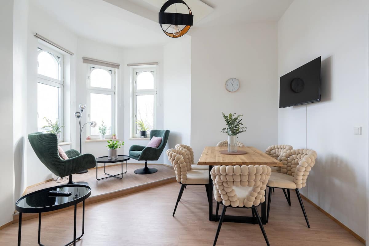 Ein modernes Wohnzimmer mit einem Holzesstisch, grünen und beigen Stühlen, einem an der Wand montierten Fernseher und großen Fenstern.