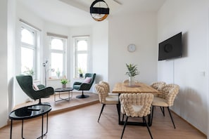 Ein modernes Wohnzimmer mit einem Holzesstisch, grünen und beigen Stühlen, einem an der Wand montierten Fernseher und großen Fenstern.
