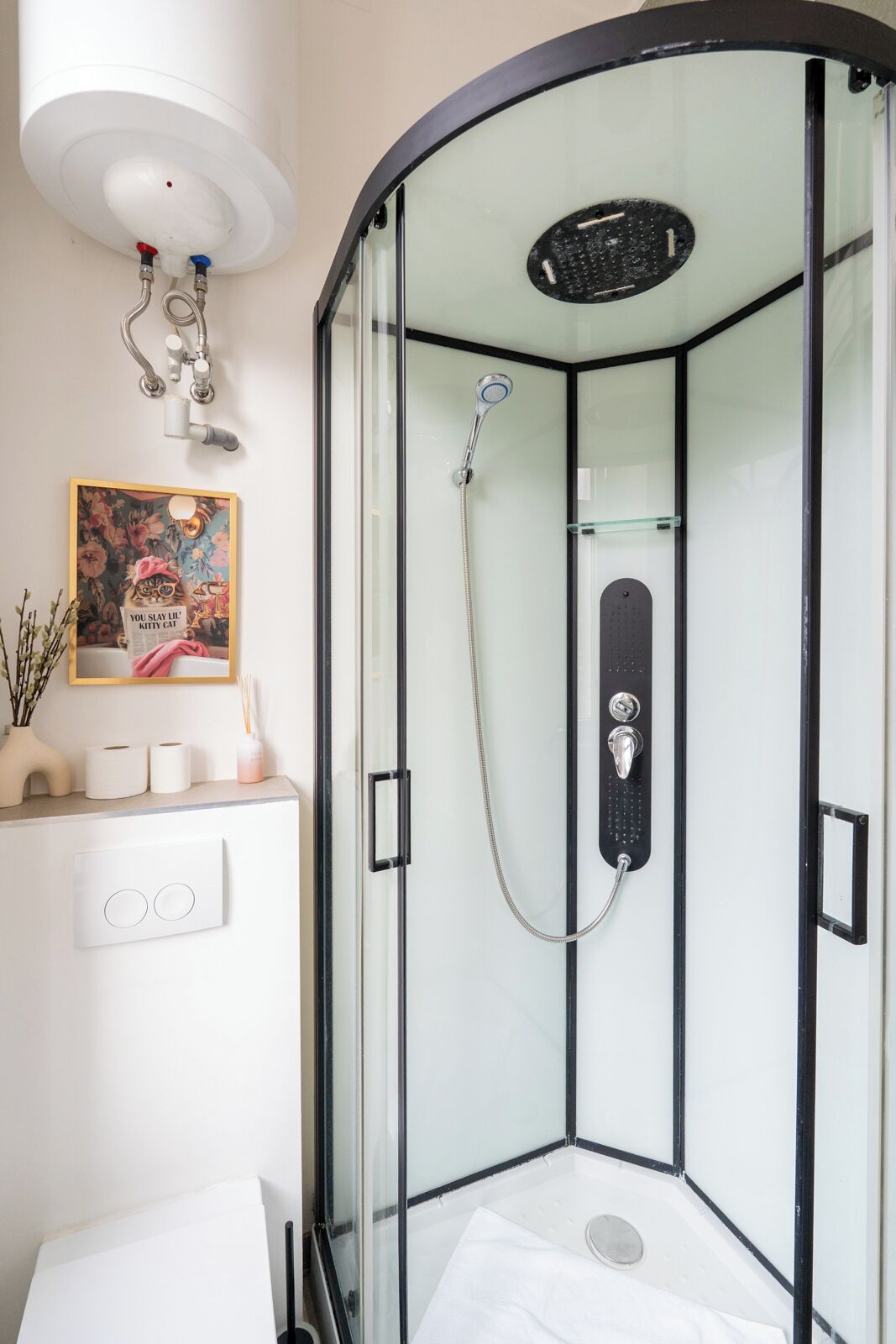 Une douche fermée par des parois en verre, dotée d’un pommeau de douche à main, d’un panneau de commande et d’un tableau encadré au mur.
