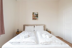 A double bed with white linens, two bedside tables with lamps, a wall-mounted painting, and a white door.