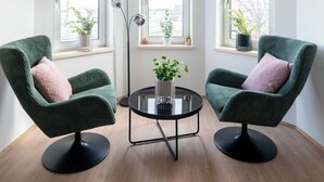 Une pièce avec deux fauteuils verts, une petite table noire ornée d’une plante et trois fenêtres à cadre blanc.