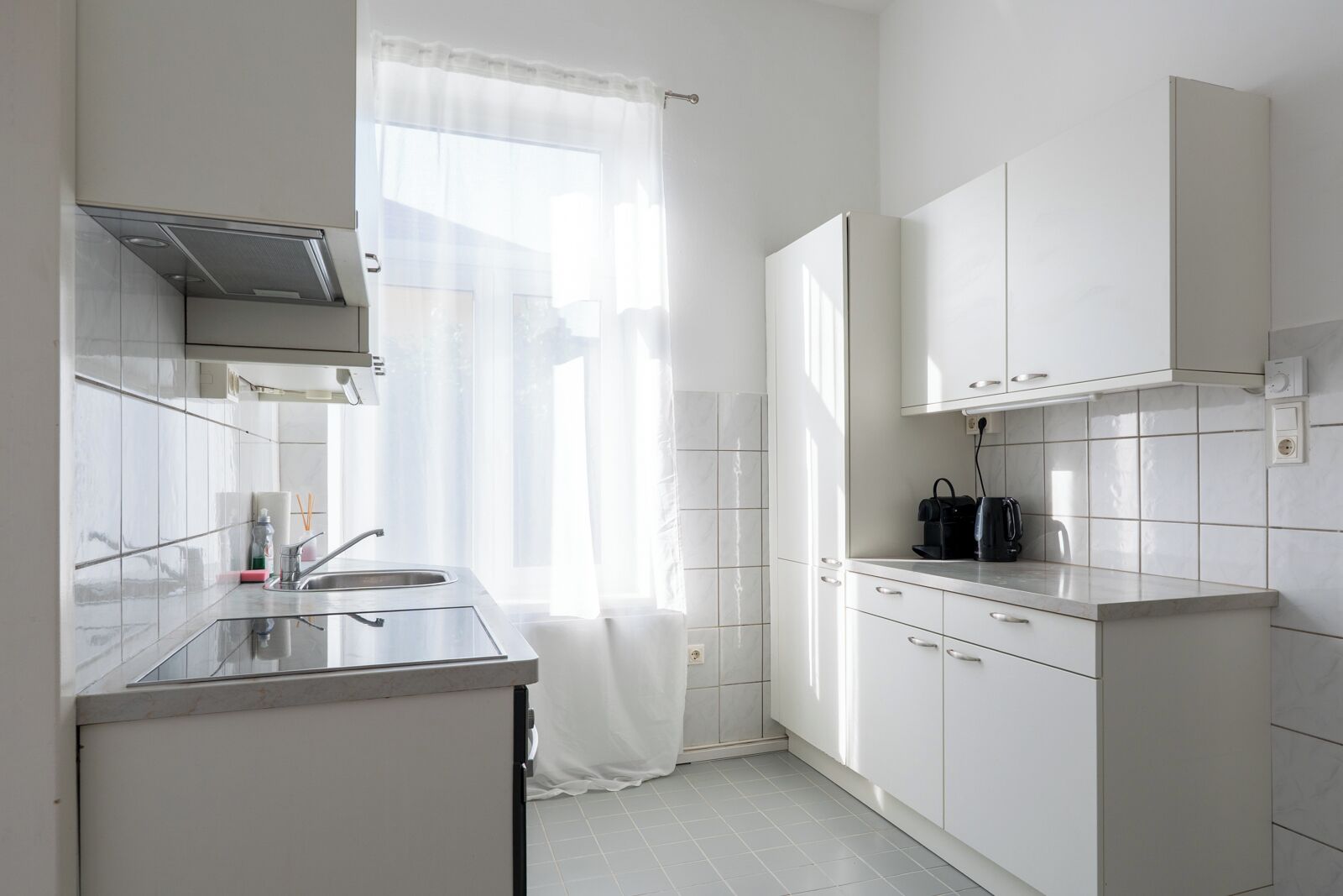 Una cocina moderna con armarios blancos, un lavamanos de acero inoxidable y una ventana con cortinas translúcidas.