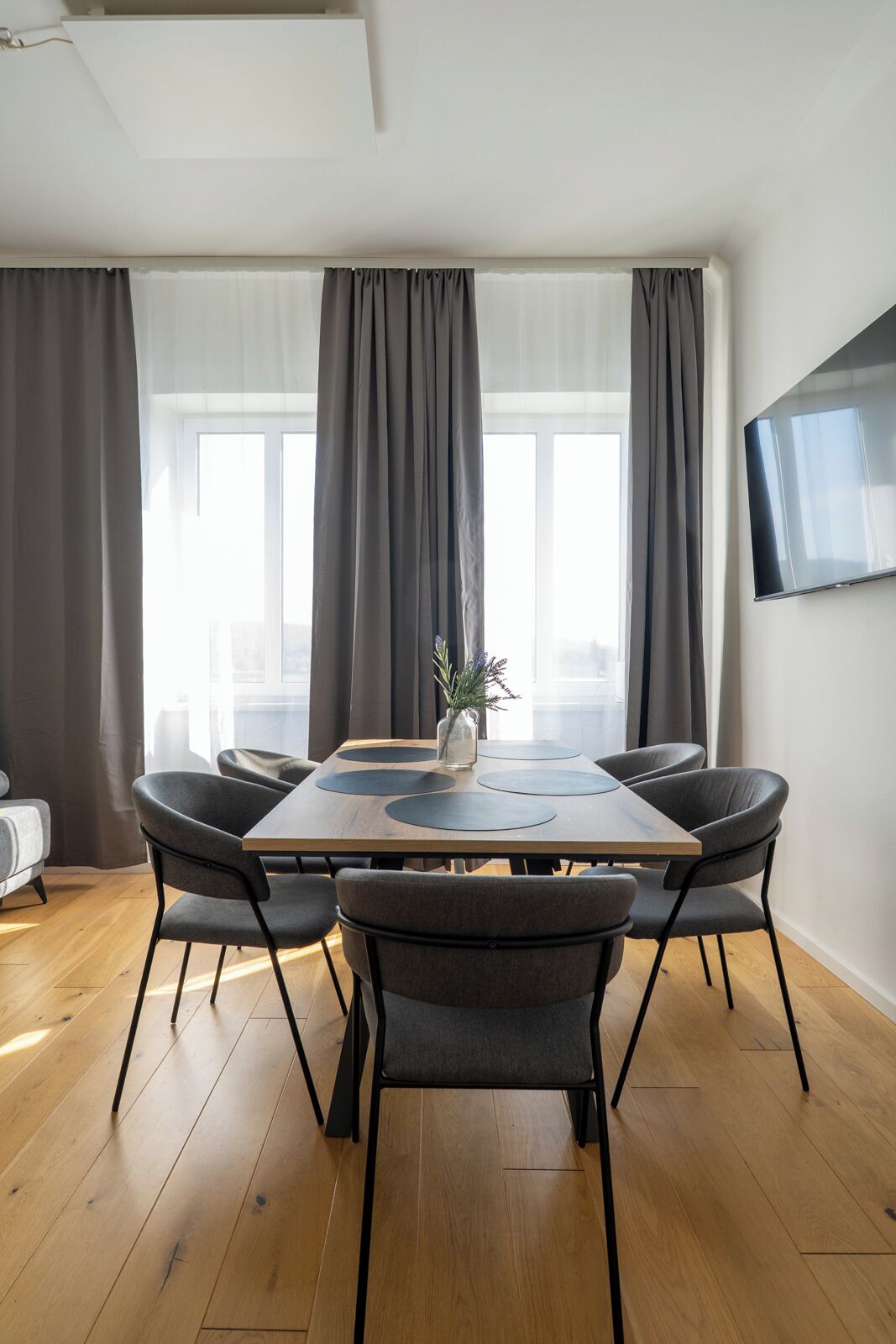 Ein moderner Essbereich mit einem Holztisch, sechs grauen Stühlen und einem großen Fenster mit grauen Vorhängen.