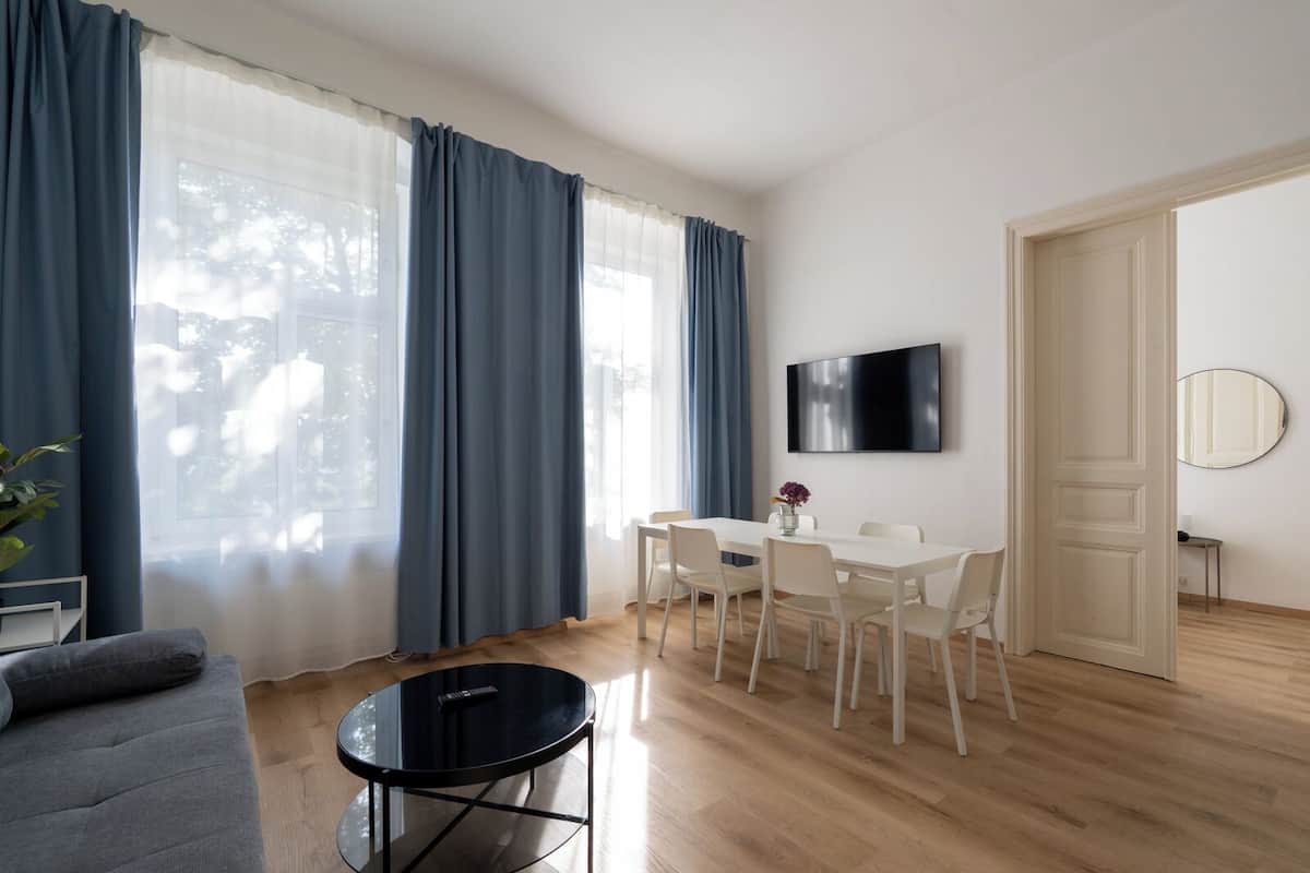 Ein modernes Wohnzimmer mit einer Couch, einem runden schwarzen Tisch, einem Essbereich mit Stühlen, einem an der Wand montierten Fernseher und einer Tür zu einem anderen Raum.