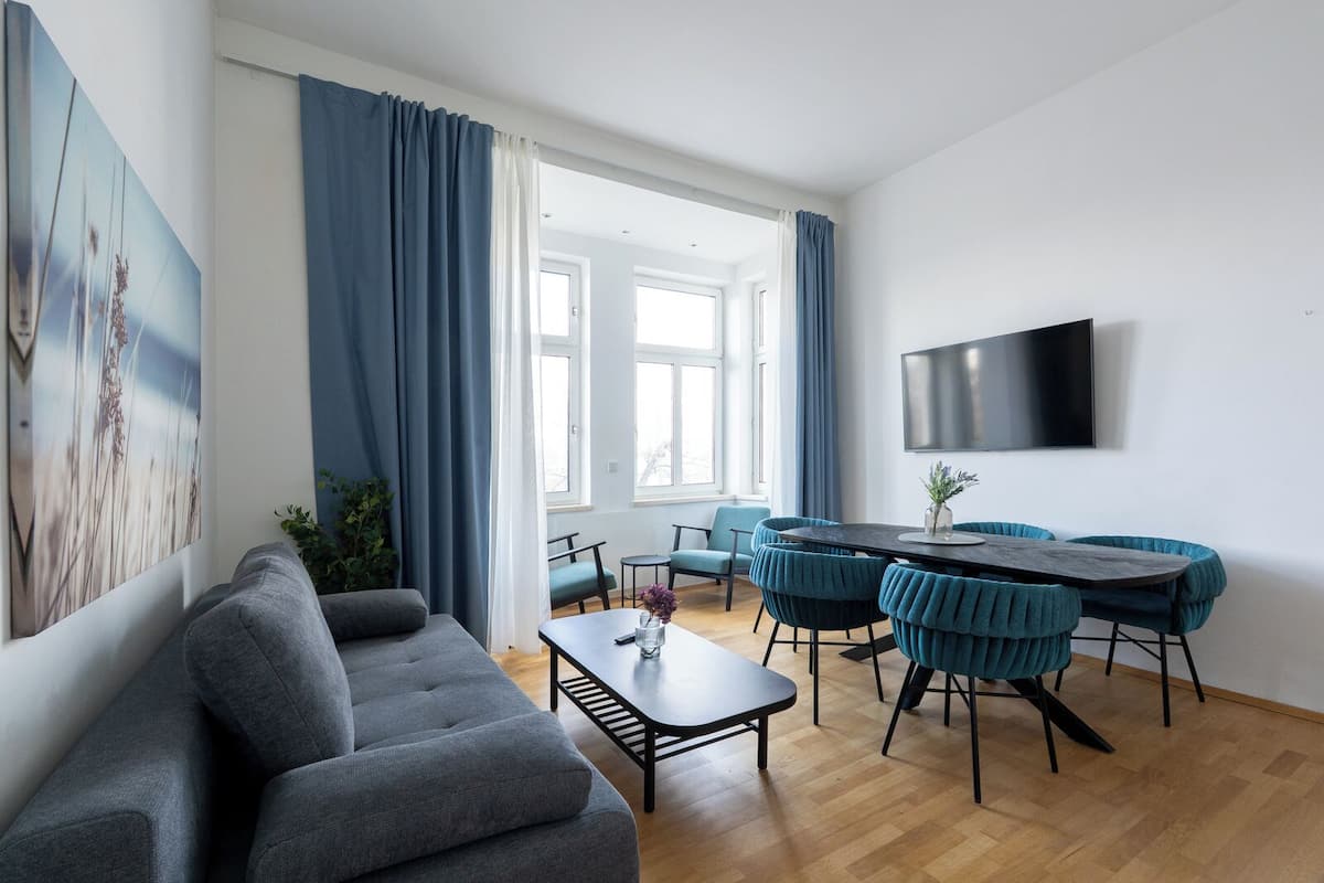 Ein modernes Wohnzimmer mit einem grauen Sofa, einem Esstisch mit türkisfarbenen Stühlen, einem Flachbildfernseher an der Wand und großen Fenstern mit blauen Vorhängen.