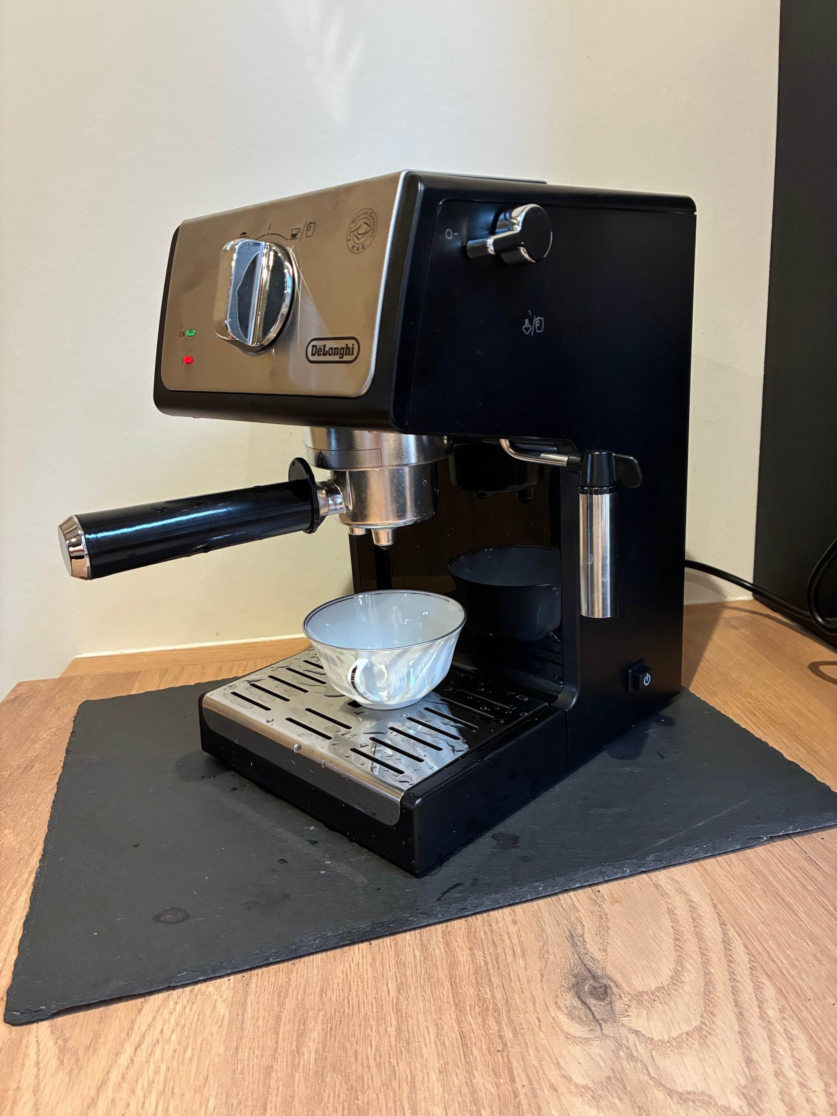 En Delonghi espressomaskine med digitalt display, en vandbeholder og en drypbakke på en sort måtte.