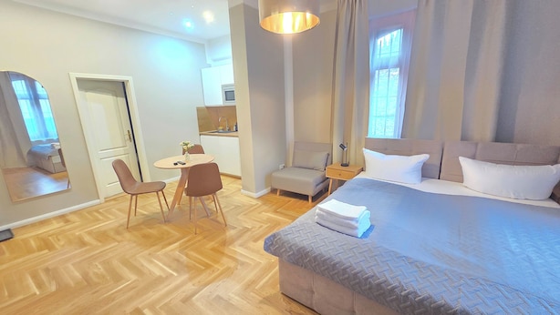 Una habitación de hotel moderna con una cama grande, una zona de comedor con mesa y sillas, una kitchenette pequeña y un espejo amplio.