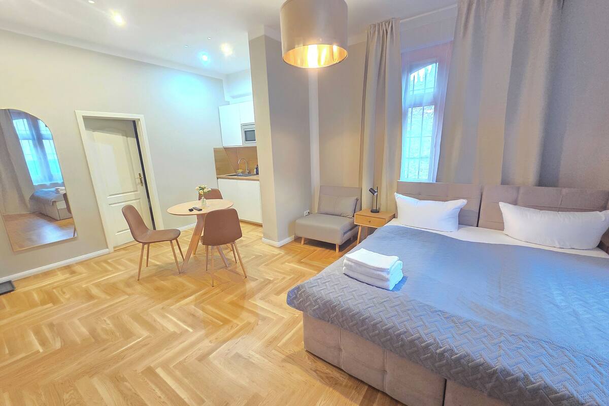 Una habitación de hotel moderna con una cama grande, una zona de comedor con mesa y sillas, una kitchenette pequeña y un espejo amplio.