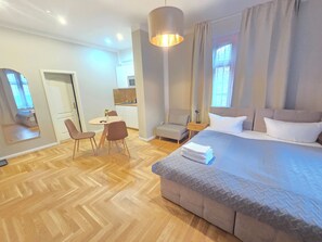 Quarto de hotel moderno com uma cama grande, área de jantar com mesa e cadeiras, pequena cozinha e um espelho amplo.