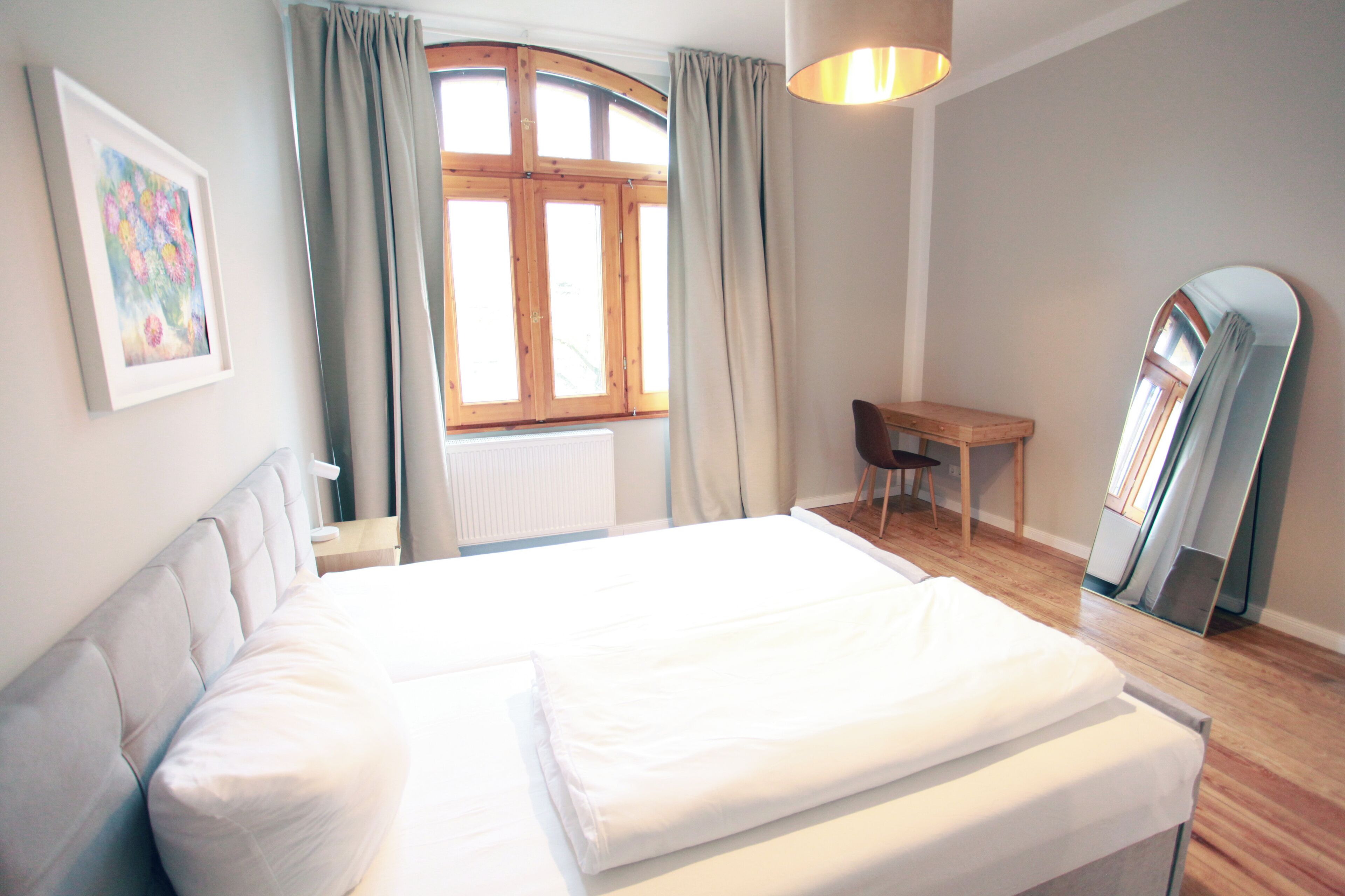 A bedroom with a large bed, a desk, a chair, a window with curtains, and a mirror.