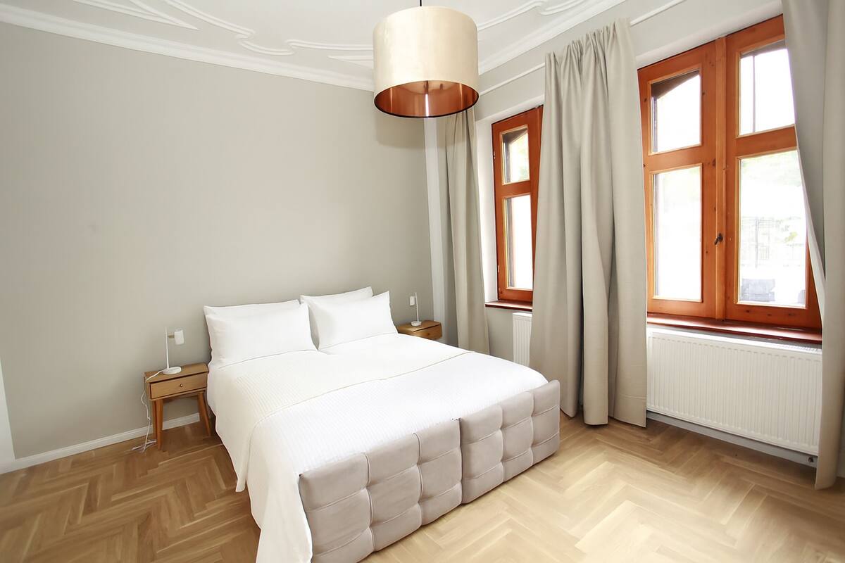 Un dormitorio con una cama grande, una mesita de noche, una lámpara y una ventana con cortinas.
