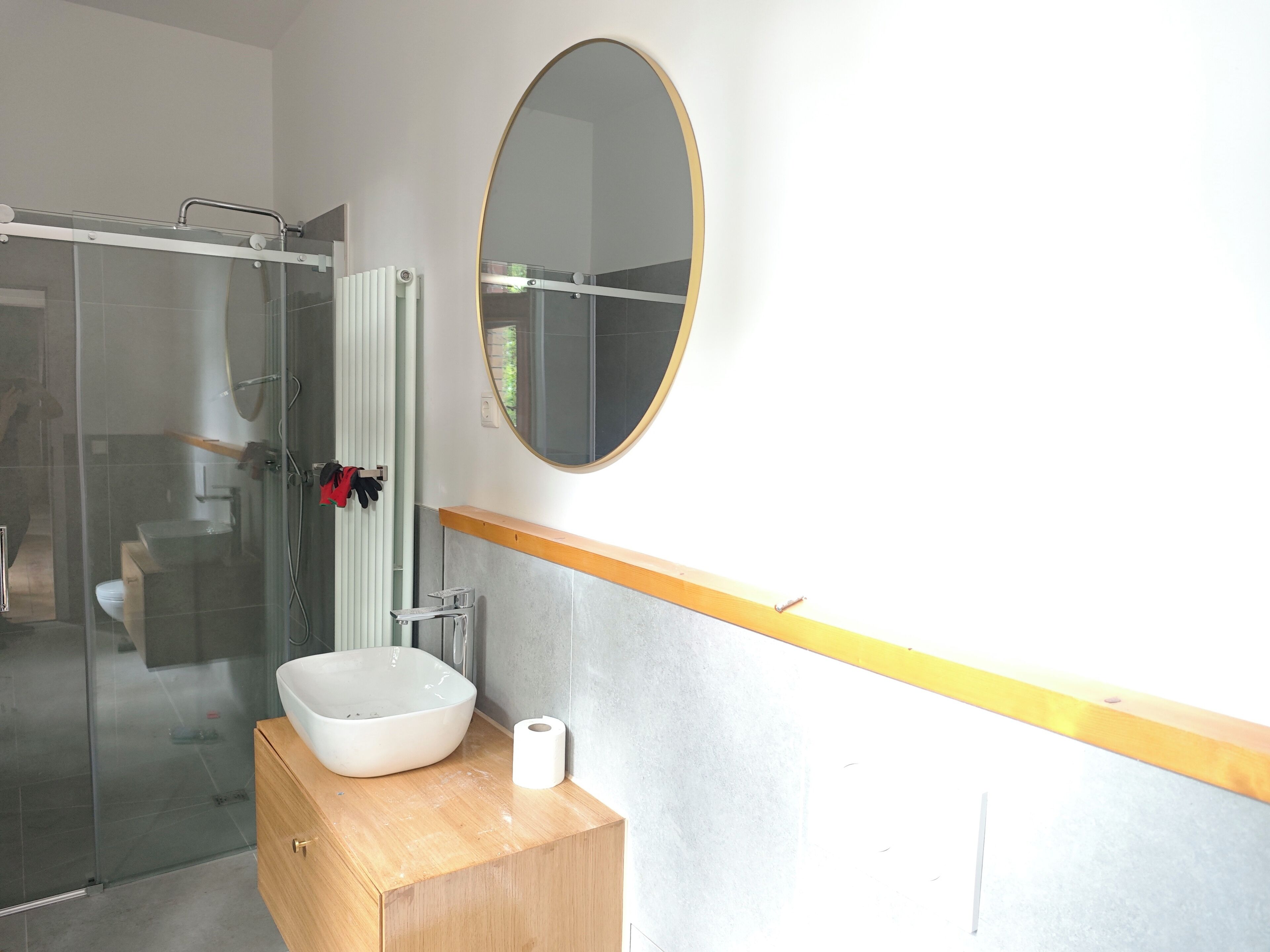 Un baño con cabina de ducha de vidrio, un lavamanos blanco con tocador de madera, un espejo redondo y un inodoro.