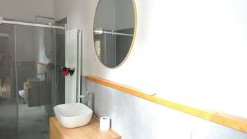 Un baño con cabina de ducha de vidrio, un lavamanos blanco con tocador de madera, un espejo redondo y un inodoro.