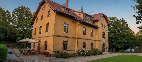 A two-story brick building with a sloped roof, multiple windows, and a small garden area with a table and chairs. -  Alter Bahnhof Stötteritz (Leipzig)