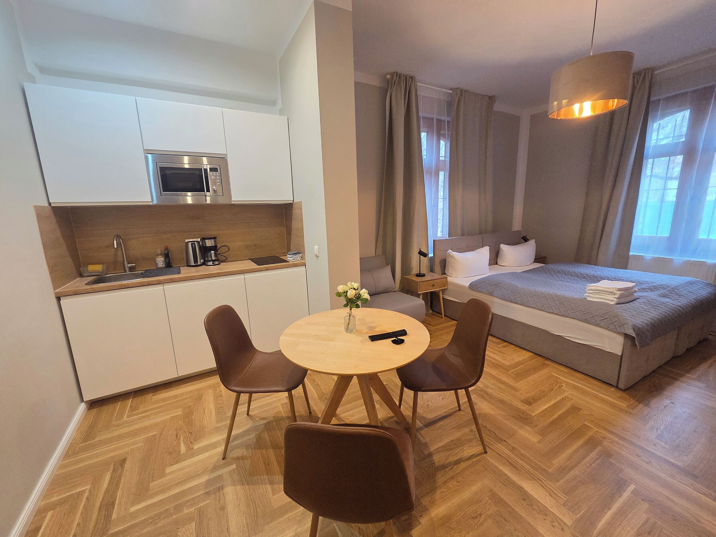 A compact hotel room with a kitchenette, a bed, a dining area with a table and chairs, and a large window.