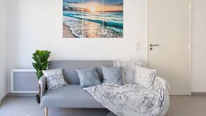 Ein Zimmer mit grauem Sofa, einem Topf mit Pflanze und einem Wandbild mit Sonnenuntergang.