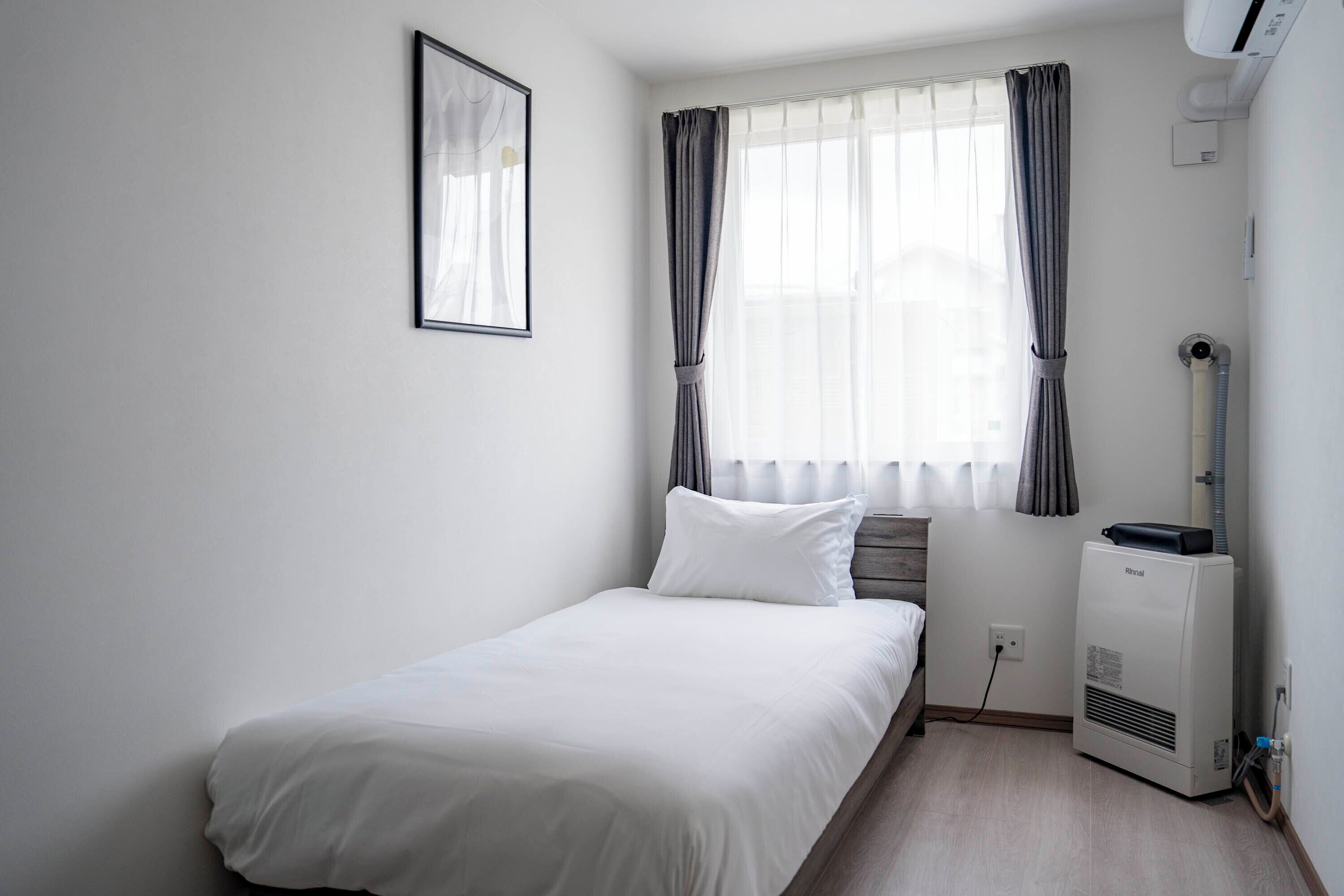 A small, neatly arranged bedroom with a single bed, a window with curtains, a wall-mounted air conditioner, and a framed picture.