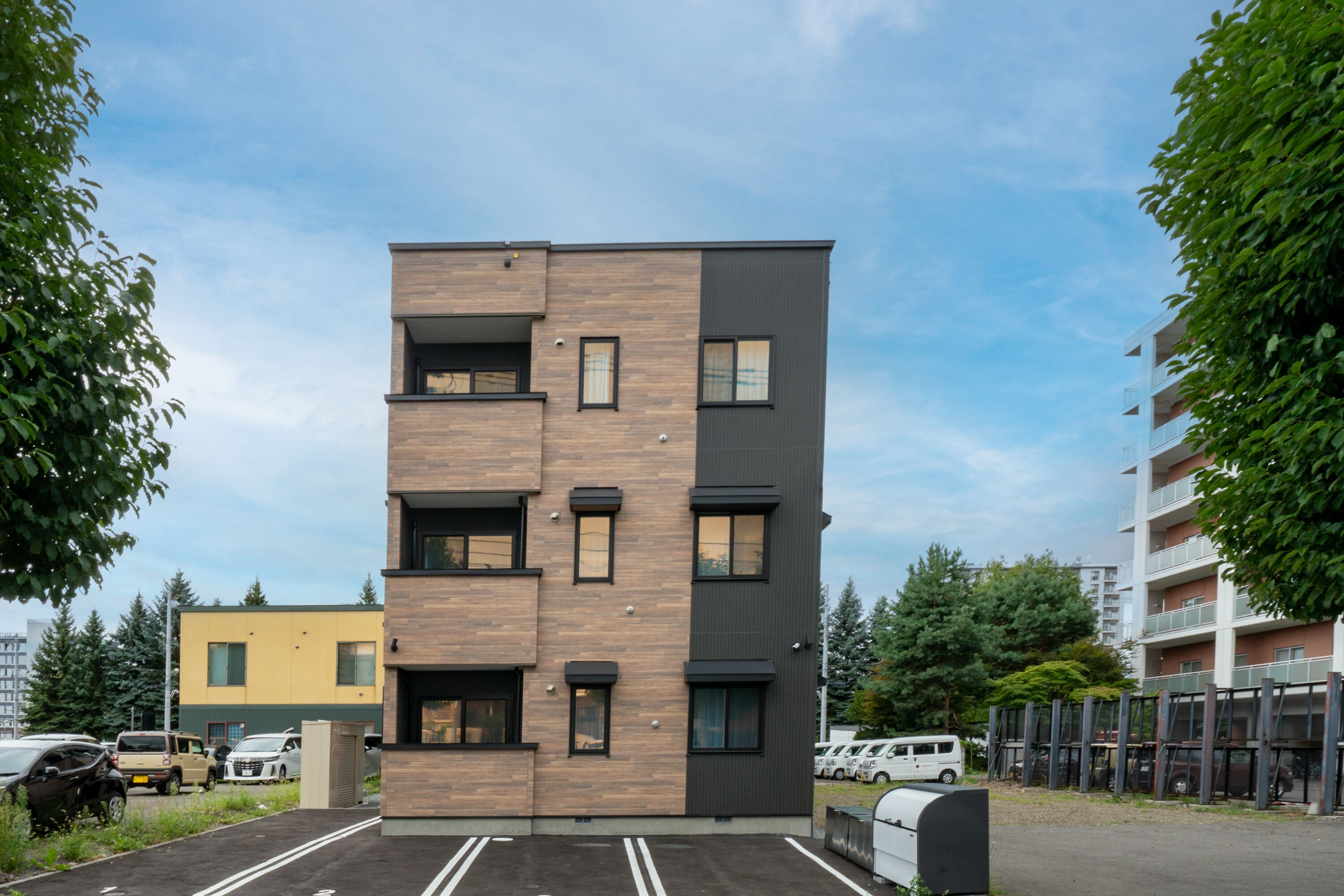 A multi-story building with a modern design, featuring a mix of wood and metal panels, multiple balconies, and a parking area with several cars.