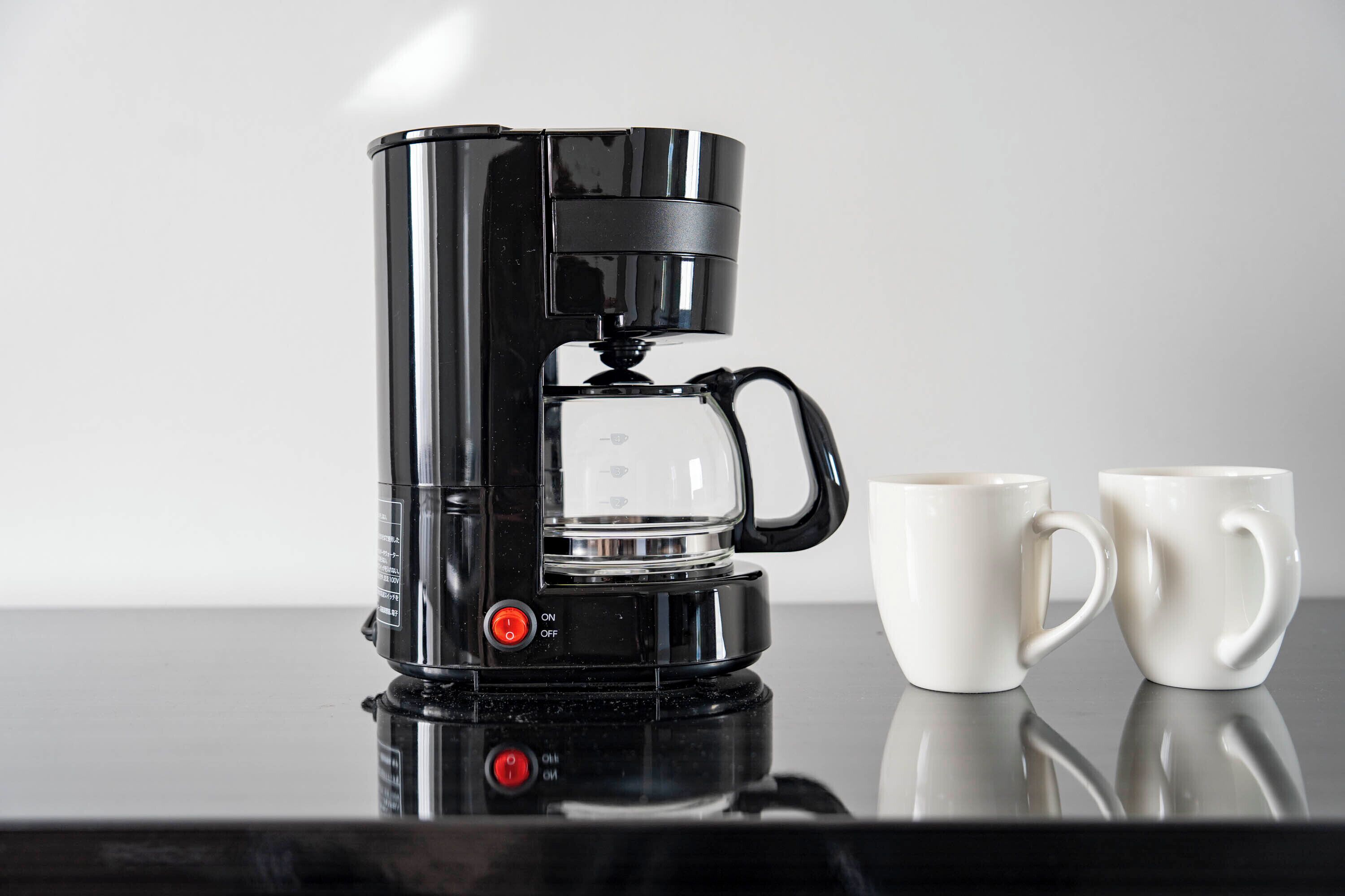 A black coffee maker with two white mugs on a reflective surface.