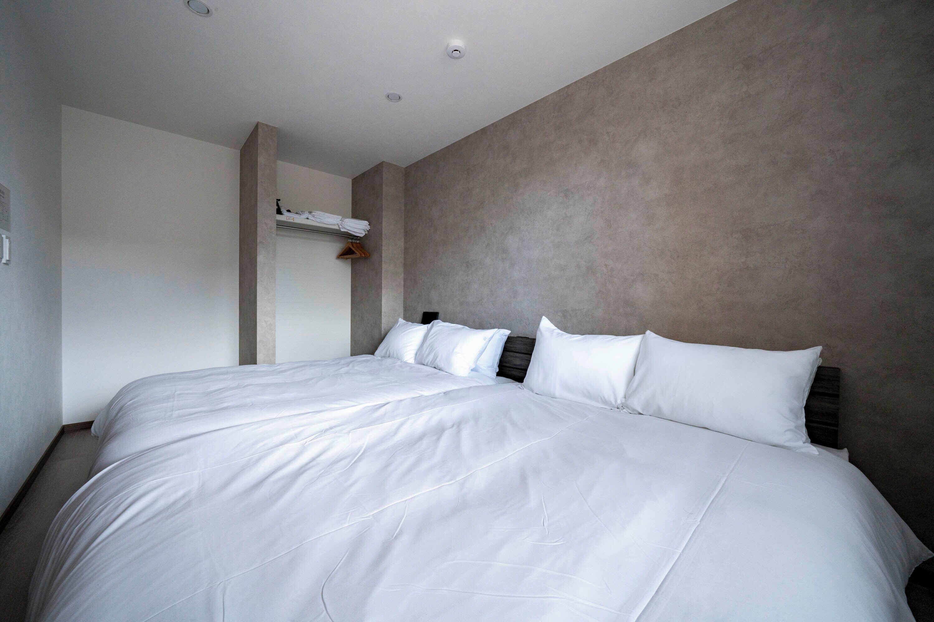 A large bed with white linens in a modern room with a concrete accent wall and a small shelf with towels.
