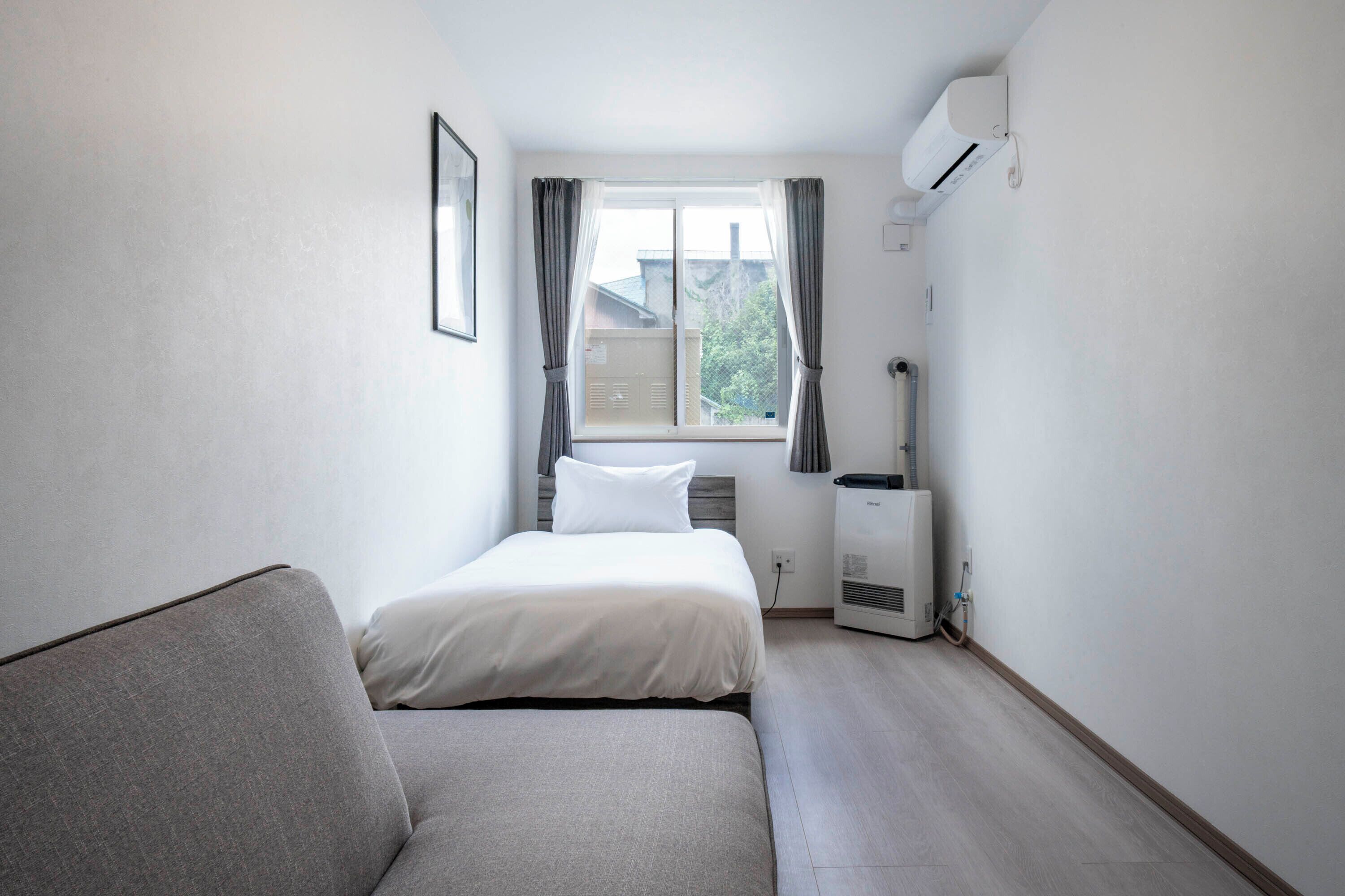 A small, clean room with a bed, a sofa, a window with curtains, an air conditioner, and a heater.