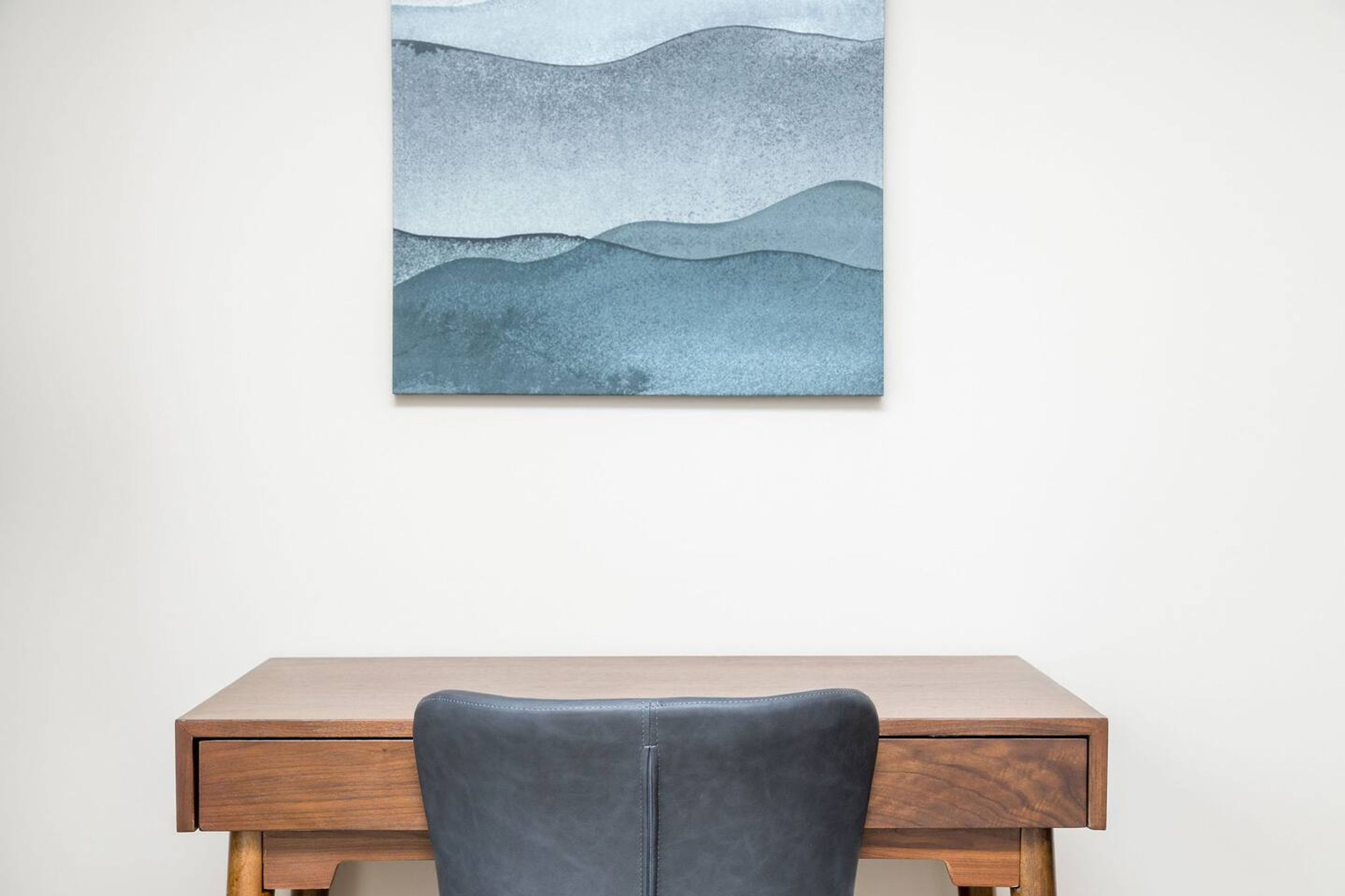 A wooden desk with a leather chair and a wall-mounted abstract painting.