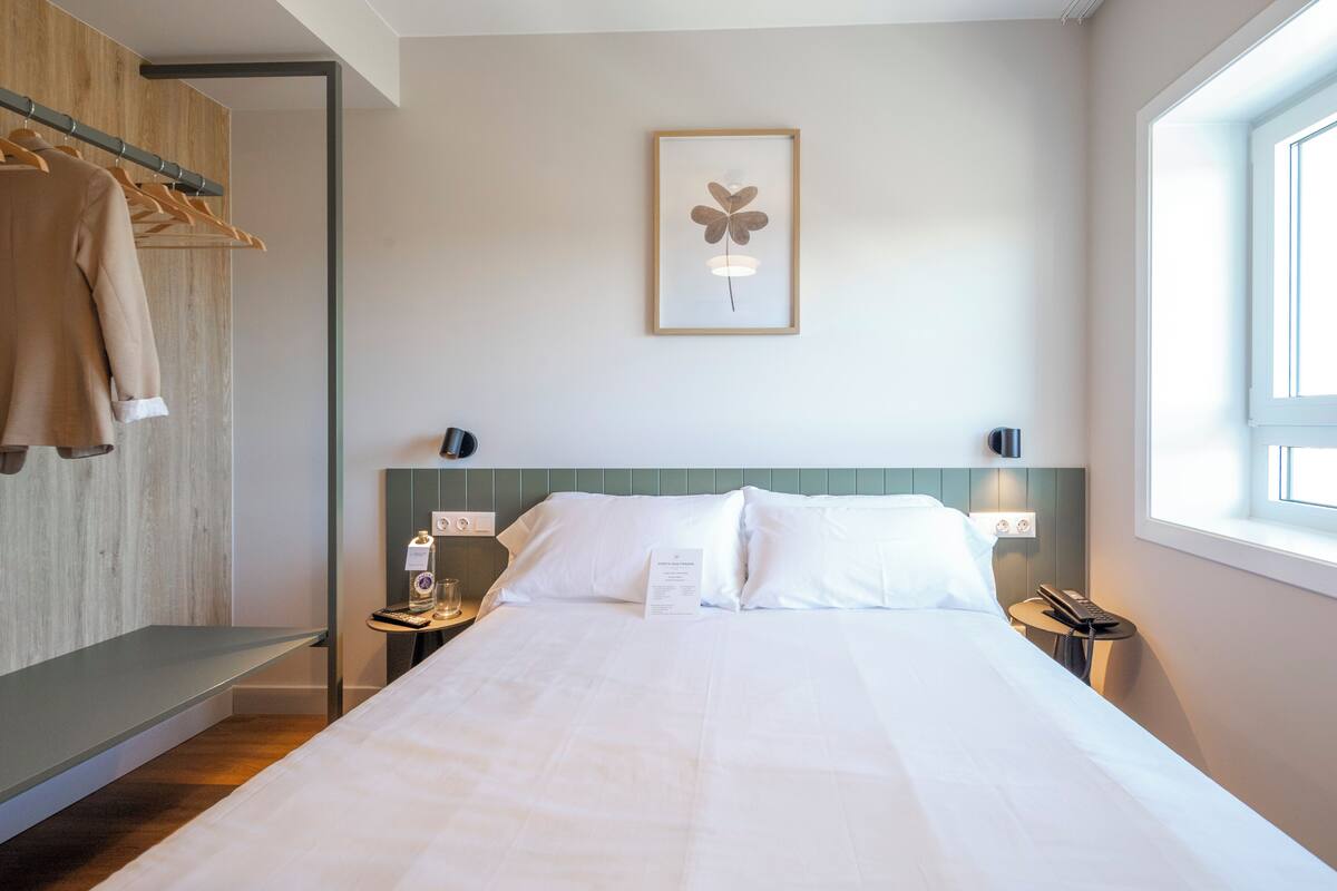 Ein Hotelzimmer mit einem Bett, einem Nachttisch mit Flasche und Glas, einem gerahmten Bild an der Wand und einem Fenster.