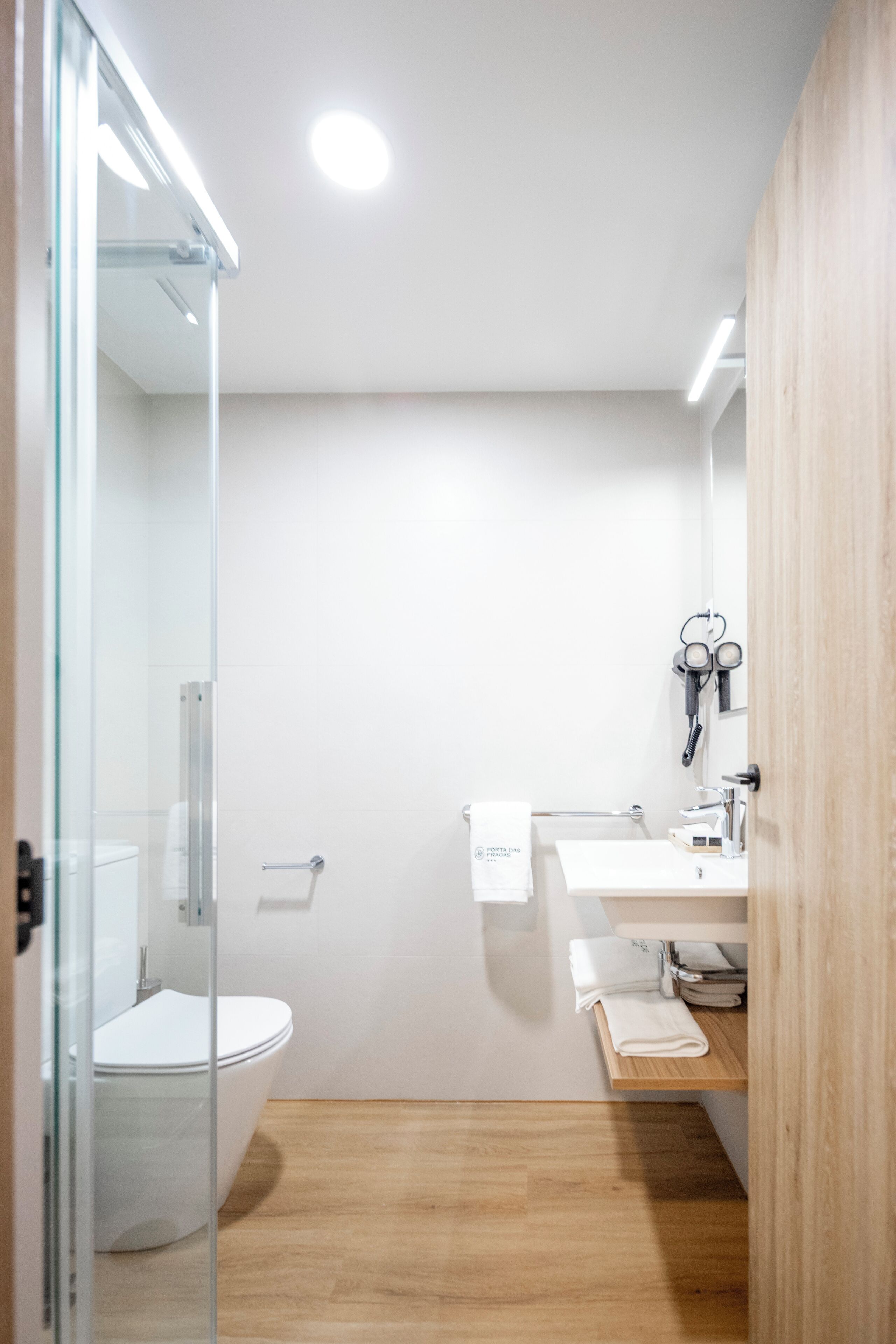 A modern bathroom with a glass shower enclosure, a white toilet, a sink with a towel rack, and a wooden floor.