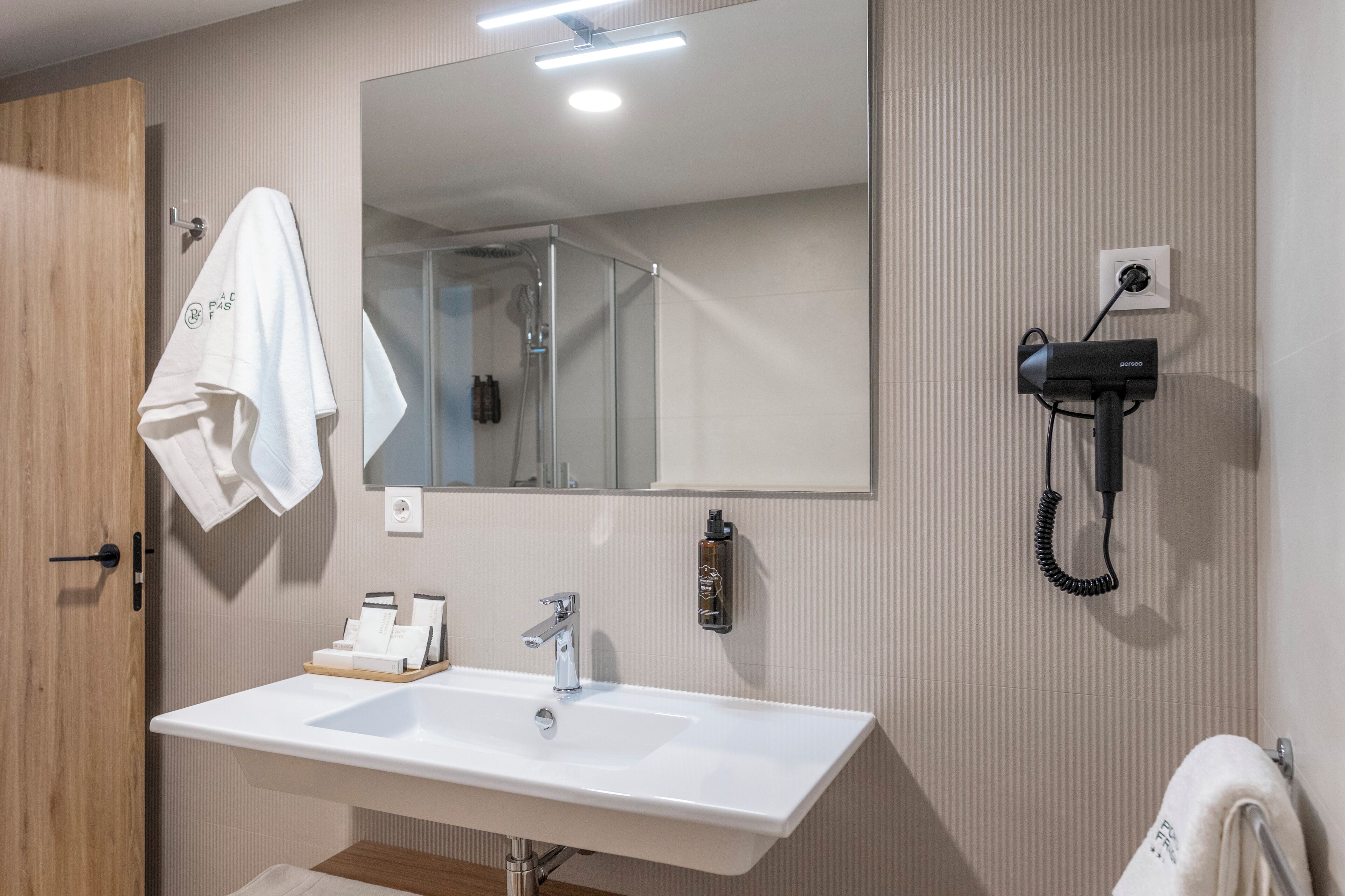 Un baño con un lavamanos blanco, un espejo, toallero, secador de pelo y una botella de jabón para manos.