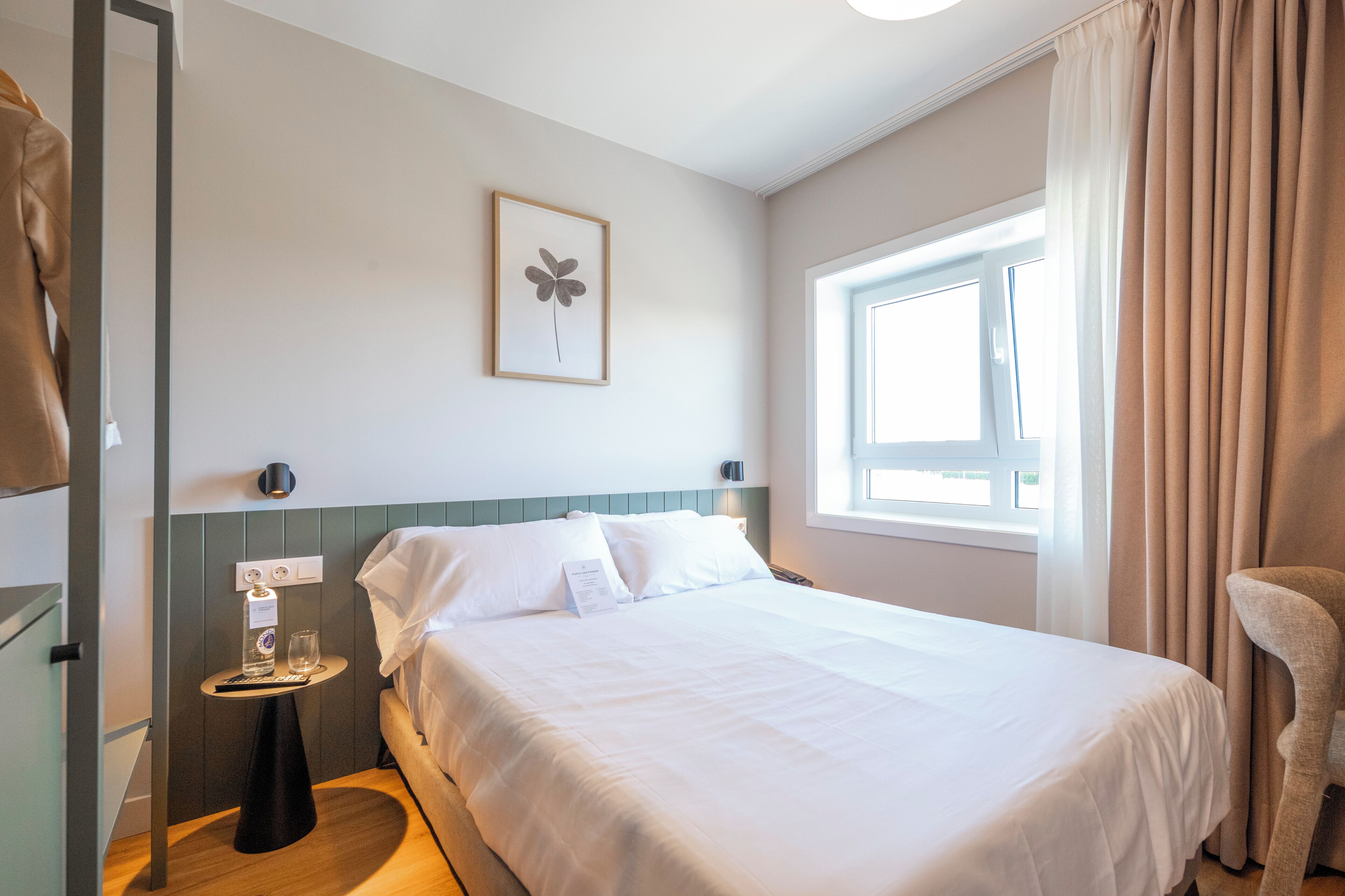 Une chambre d’hôtel avec un lit, une table de chevet sur laquelle se trouvent une bouteille et un verre, un tableau encadré au mur et une fenêtre avec des rideaux.