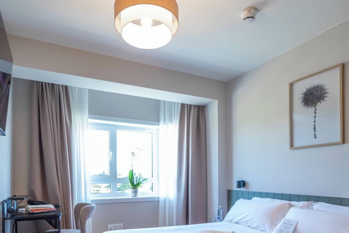 Ein Hotelzimmer mit Bett, Schreibtisch, Stuhl, einem Fenster mit Vorhängen und einer Deckenleuchte.