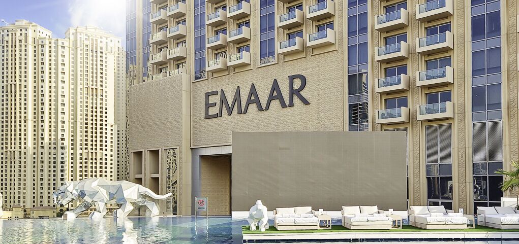 Een hotel met een zwembadgedeelte, een groot bord met de tekst 'EMAAR' en een modern gebouw met balkons op de achtergrond.