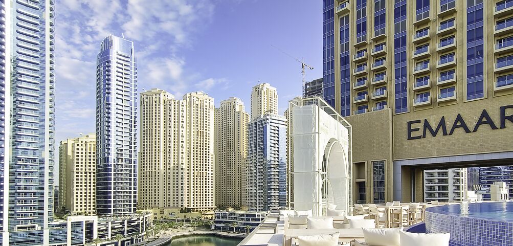 A high-rise building with multiple floors, a swimming pool area with lounge chairs, and a clear sky.