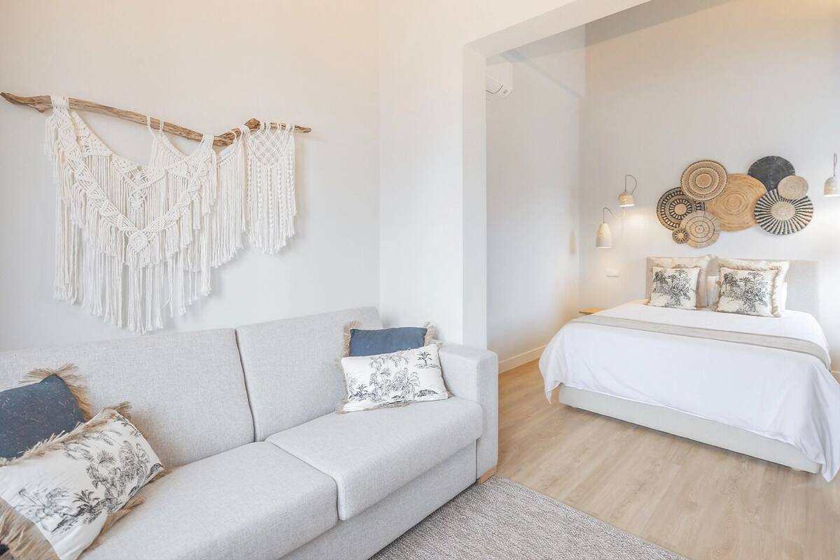 A modern bedroom with a grey sofa, a white bed, macramé wall hangings, and decorative wall art.