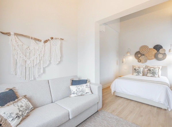 A modern bedroom with a grey sofa, a white bed, macramé wall hangings, and decorative wall art.