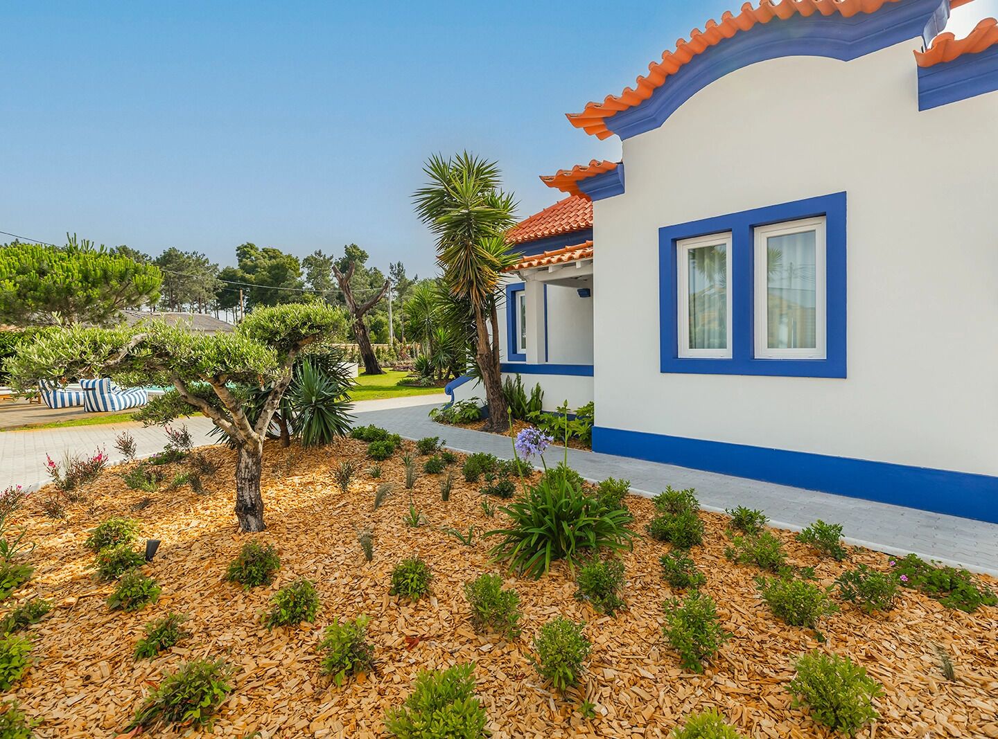 Un edificio moderno con fachada blanca y detalles azules, rodeado de un jardín ajardinado con diversas plantas y un sendero.