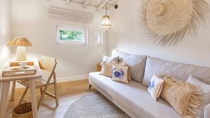 A cozy living room with a light-colored sofa, a small table with a lamp, and a wicker chair. There is a large woven wall hanging and a window with a view of greenery. - Almaa da Ria (Ilhavo)