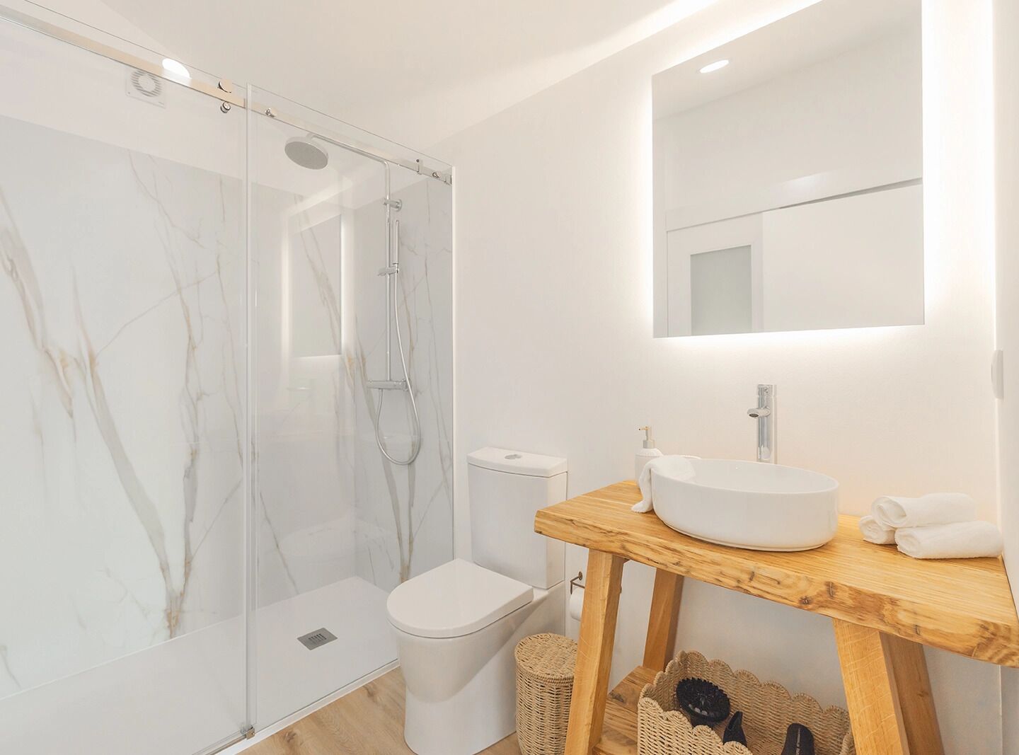 Un baño moderno con ducha envuelta en vidrio, inodoro blanco y tocador de madera con lavamanos y espejo.