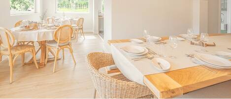 A bright dining area with wicker furniture and pendant lights, large windows offering a view of greenery.