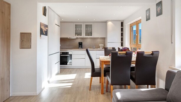 Une cuisine moderne avec des meubles blancs, un coin repas doté d’une table en bois et de chaises noires, et une vue sur l’extérieur par une fenêtre.