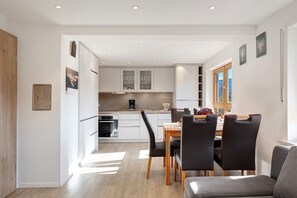 Eine moderne Küche mit weißen Schränken, ein Essbereich mit Holztisch und schwarzen Stühlen und ein Blick ins Freie durch ein Fenster.