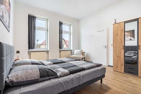 Un dormitorio con cama, dos ventanas, un armario y vistas a edificios.