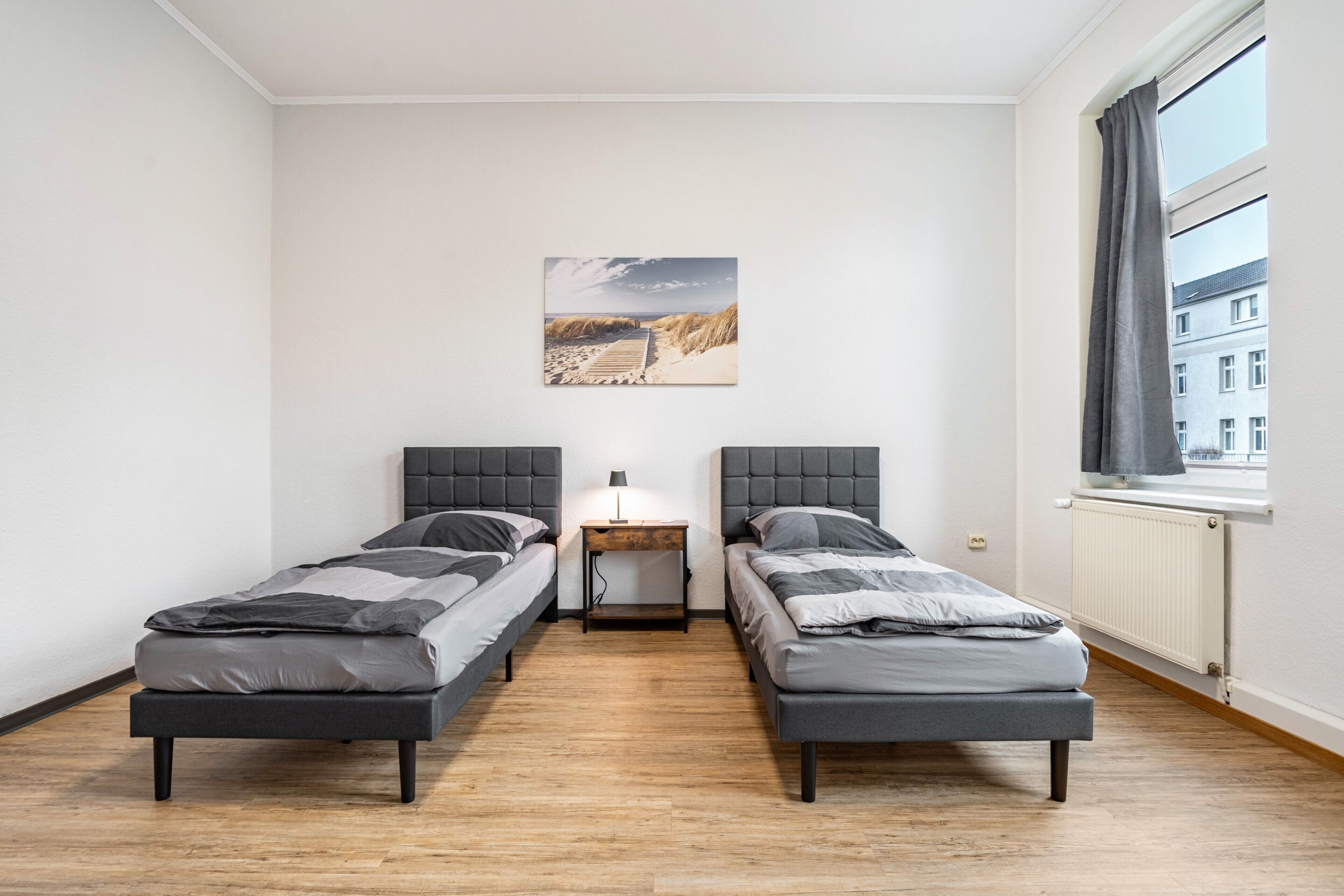 A twin room with two single beds, a nightstand, a lamp, a picture on the wall, and a window with curtains.