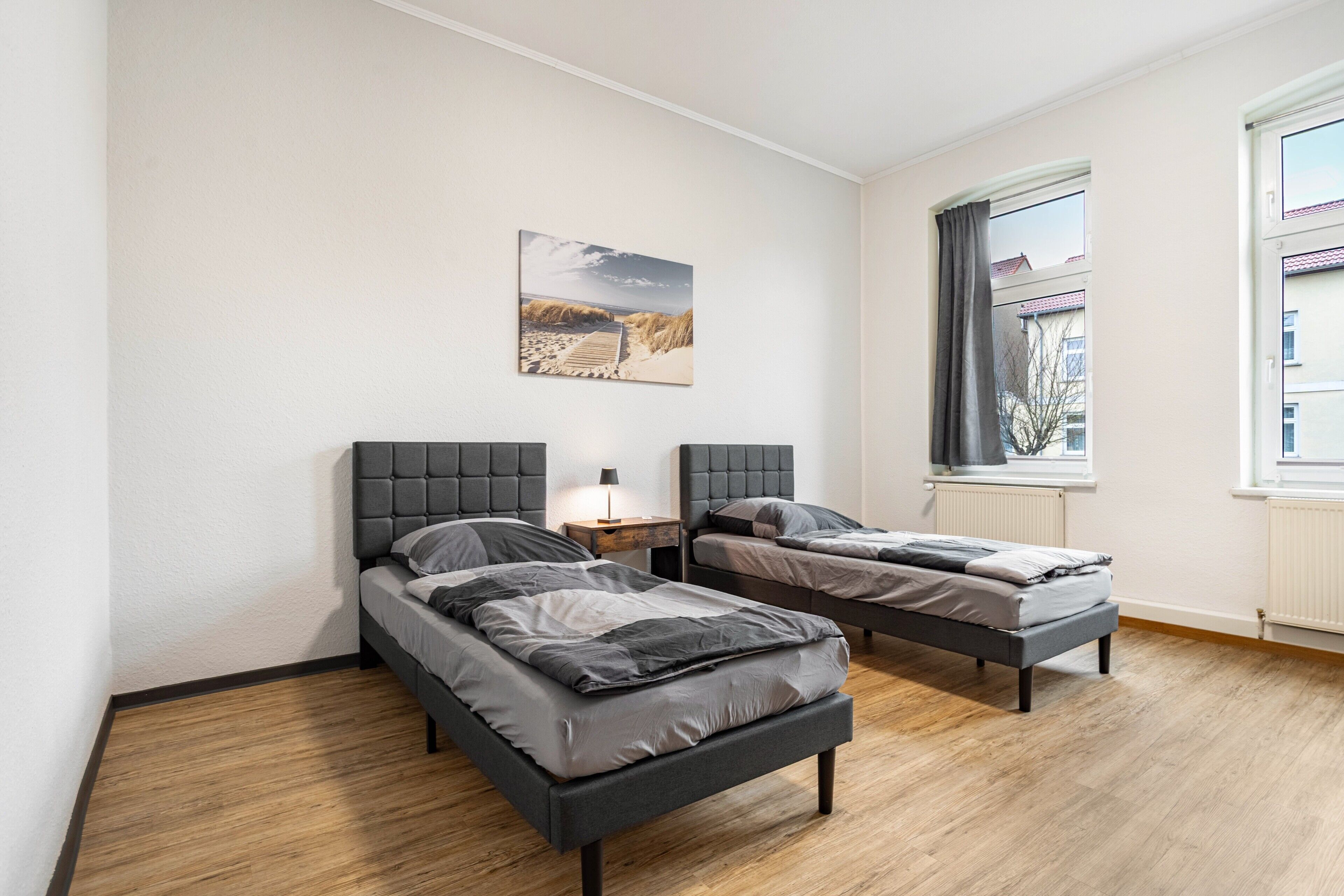 Una camera da letto con due letti, un comodino con una lampada, una finestra con tende e un quadro raffigurante una spiaggia.