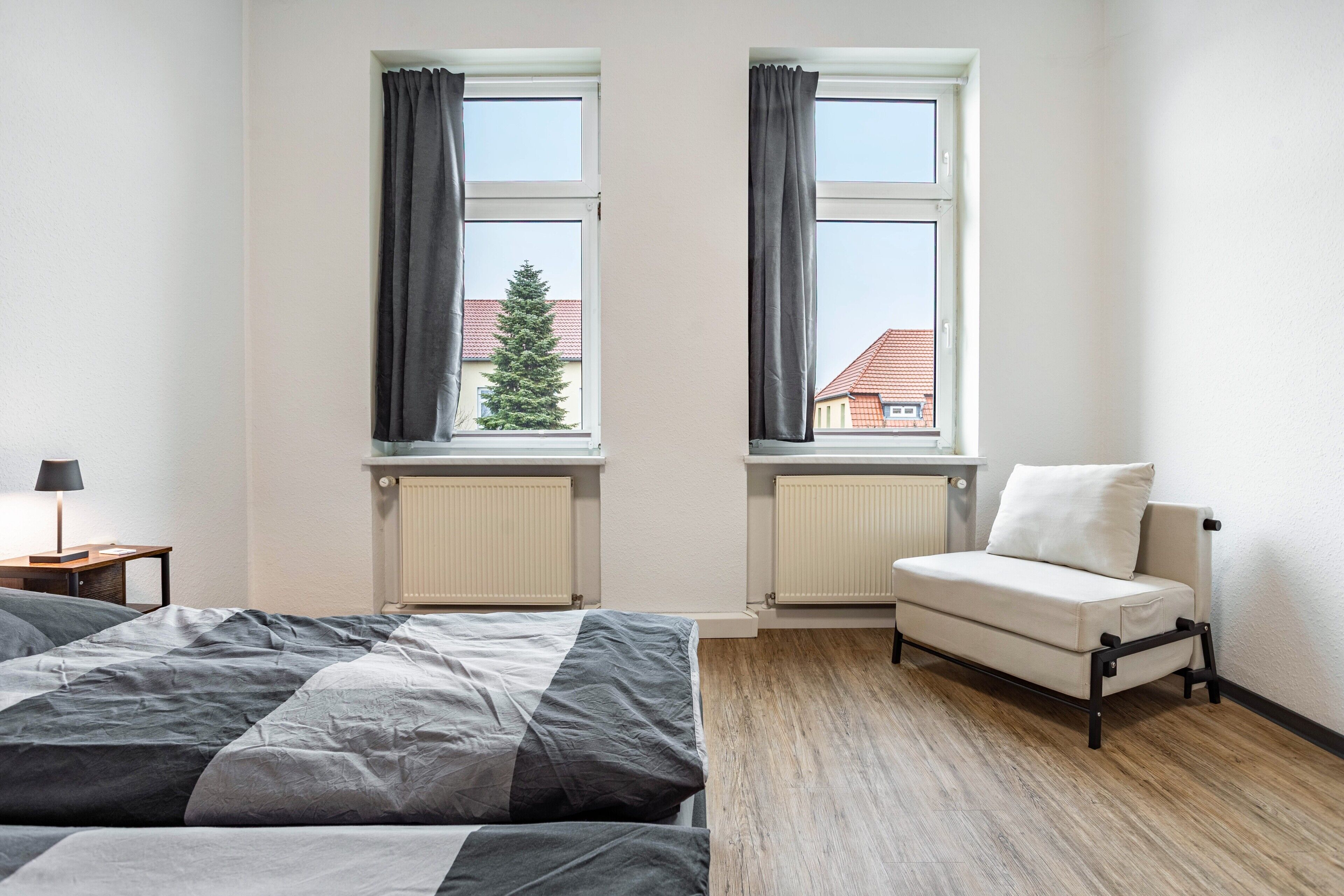 Ein Schlafzimmer mit einem Bett, einem Sessel, zwei Fenstern mit Vorhängen und einer Heizung.