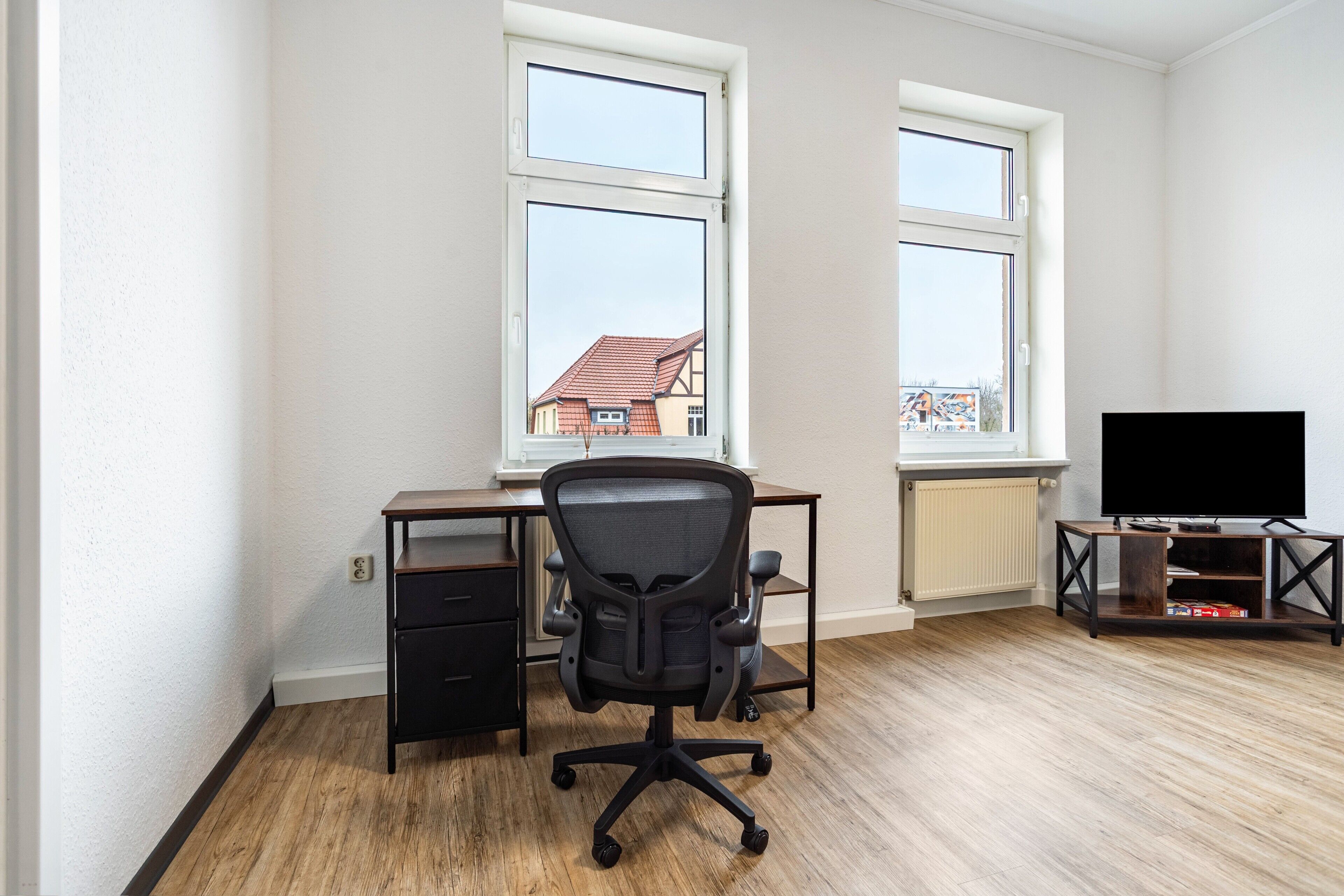 Ein Zimmer mit Schreibtisch, Bürostuhl, Fernseher und Fenstern.