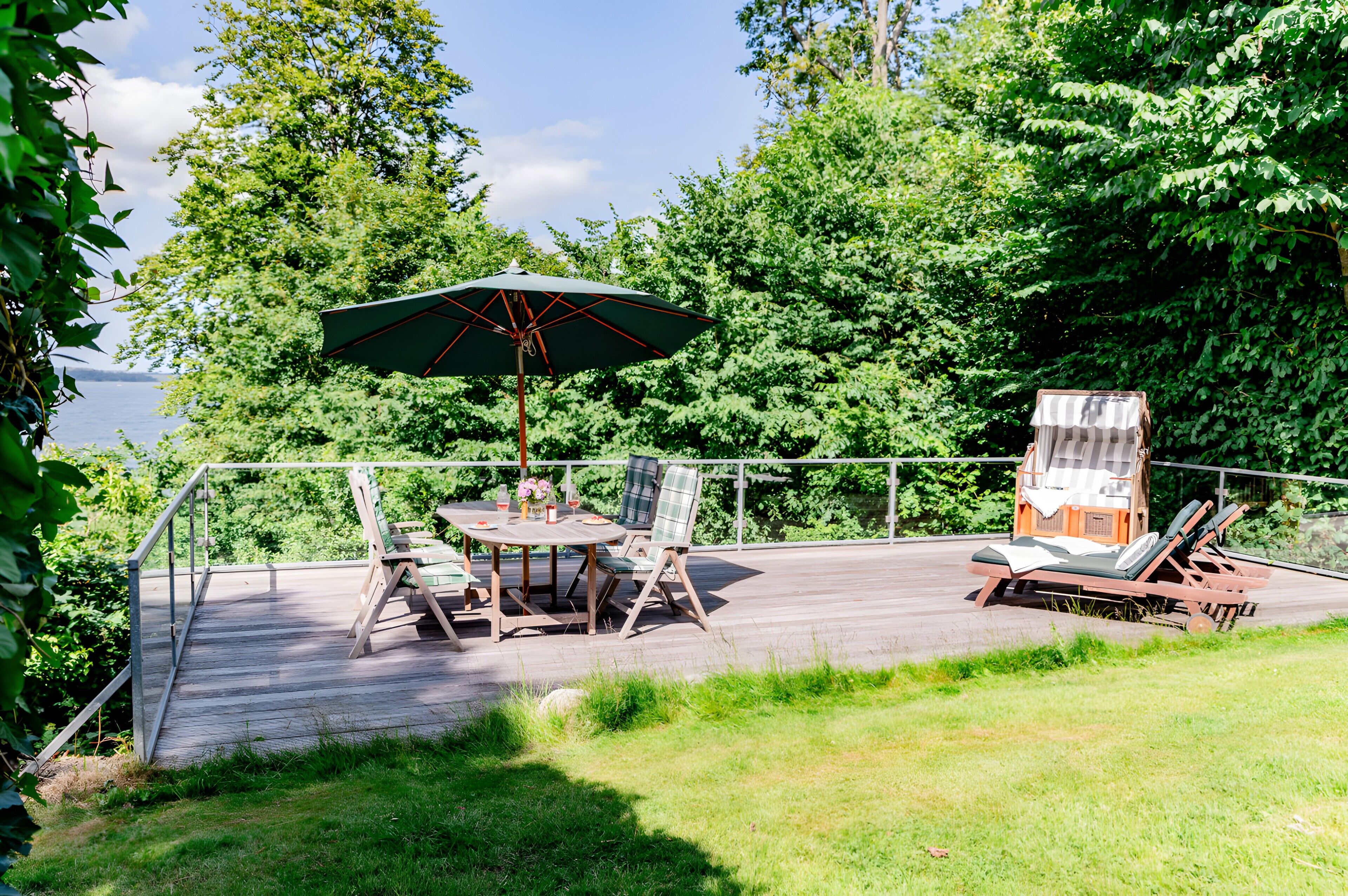 A wooden deck with a table and chairs, an umbrella, and a small cabin-like structure with a chair and a lounge chair.