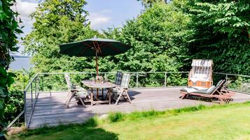 A wooden deck with a table and chairs, an umbrella, and a small cabin-like structure with a chair and a lounge chair.