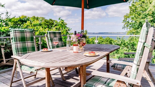 A wooden deck with a table and chairs, a table umbrella, and a scenic view. - Holiday Home 'Seeblick' with Sea View, Private Terrace and Wi-Fi (Langballig)